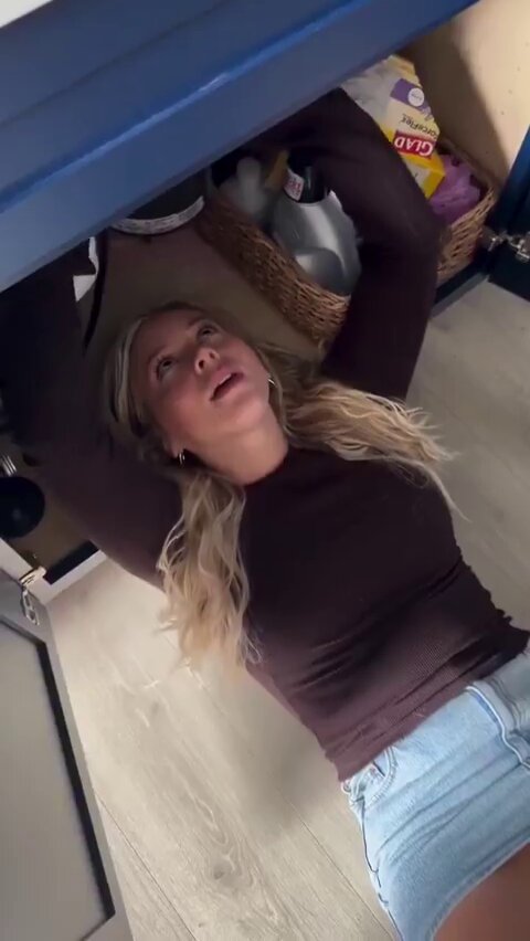 Blonde in a brown top and denim shorts looking up from under a sink